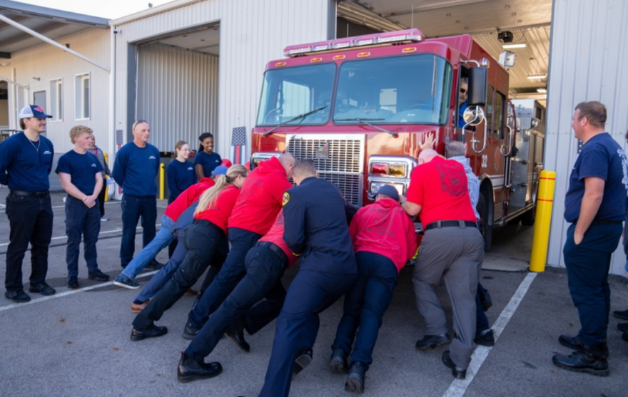 A group of around 9 people push a fire engine into its bay. Most of them are wearing red shirts. They are surrounded by onlookers wearing navy shirts. A man sits in the driver's seat of the fire engine. 