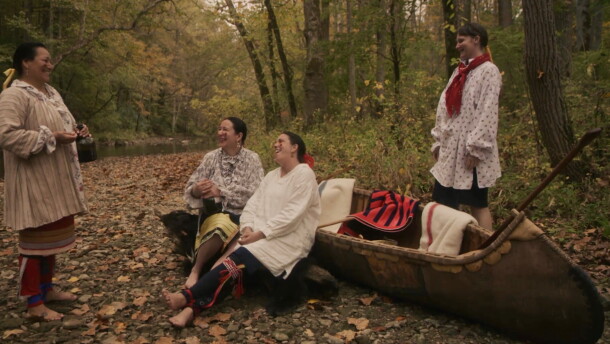 Four individuals smile and laugh while in a forest near a body of water. Two of the individuals are leaning against a boat, while the other two are standing. 