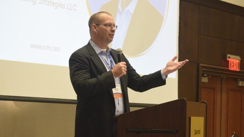 Man in a sports jacket and holding a microphone while speaking at a conference 