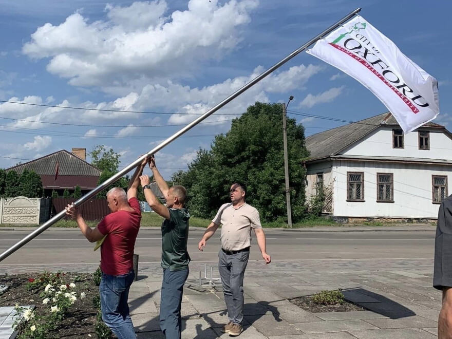 Three men raise a flag pole with a white flag and the word "Oxford" on it, in front of a residential street.