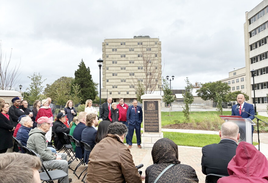 A man in a suit speaks at a podium during a dedication ceremony for a memorial plaque outdoors, with a seated audience watching.