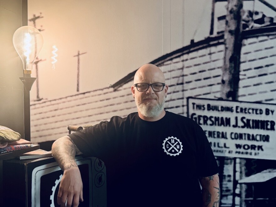 A bald man with glasses and a beard stands indoors, wearing a black T-shirt with a circular gear logo. He leans on a piece of furniture next to a retro-style exposed light bulb. A vintage sign is visible on the wall behind him.