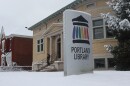 A snow-covered sign that says "Portland Library" at the front entrance of the snowy building