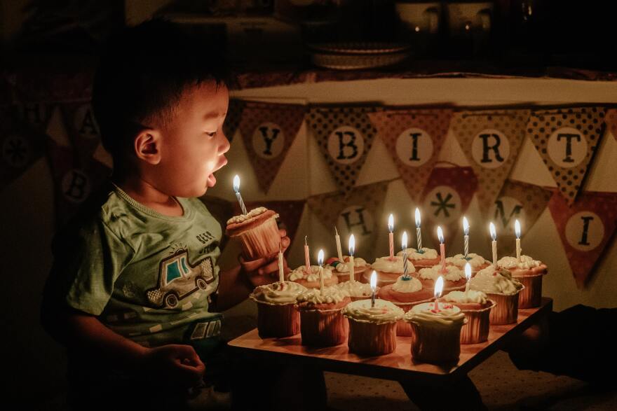 Young child of color prepares to blow out the candle in the cupcake he is holding from a trayful of cupcakes, all with lit candles