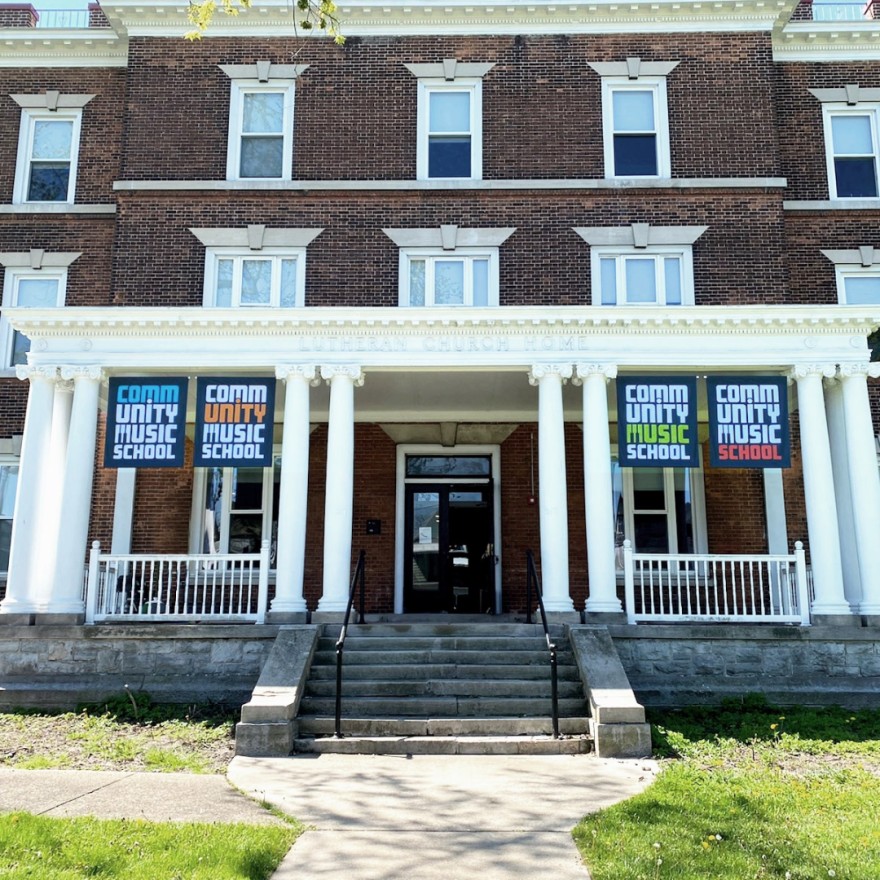 The front steps of a brick building with white pillars. There are four signs hanging from the building that read "Community Music School".