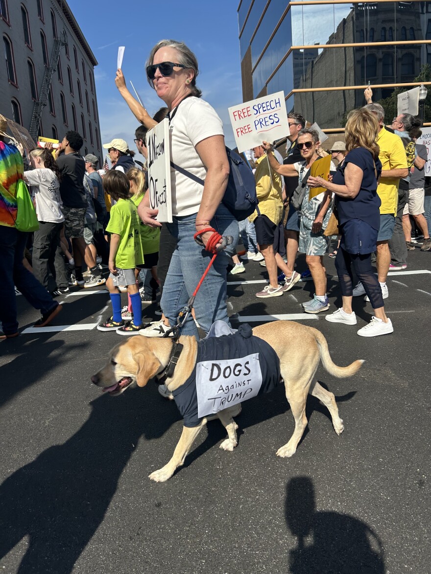 A woman walking a dog in a crowd. The dog's t-shirt says "dogs against Trump."