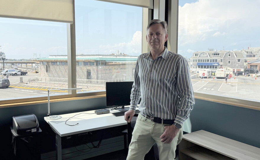 A man standing in a corner office with a view of ferry docks and seaside buildings