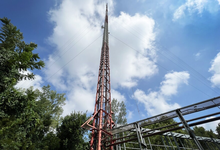 A tall, skinny steel radio tower, painted red and white is seen from below against a cloudy, blue sky. Metal framework near the base is in the foreground.