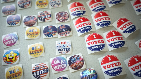 Close up image of about 30 different lapel stickers lying flat on a table. Some read "I Voted Today" with a red, white and blue design. Others read "Be a Voter" in colorful designs.