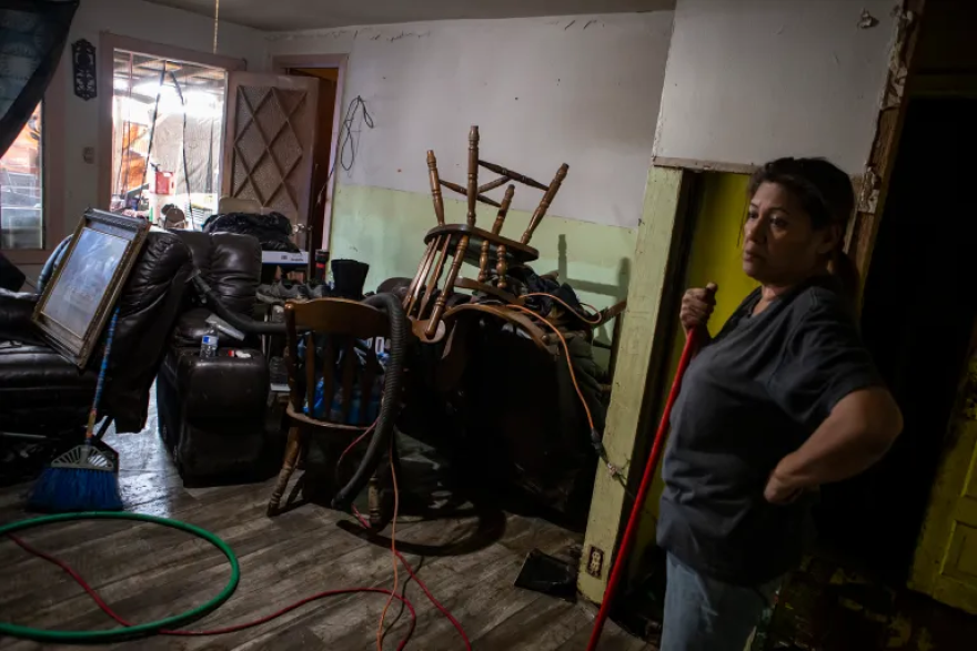 A woman standing among stacked furniture in a room with flood damage.