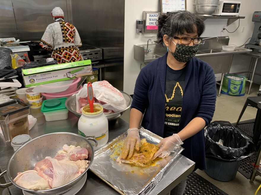 Theary Ngeth works at a stainless steel table, rubbing a spiced paste into raw chicken. She's wearing a mask and the chicken is on a tray lined with foil.