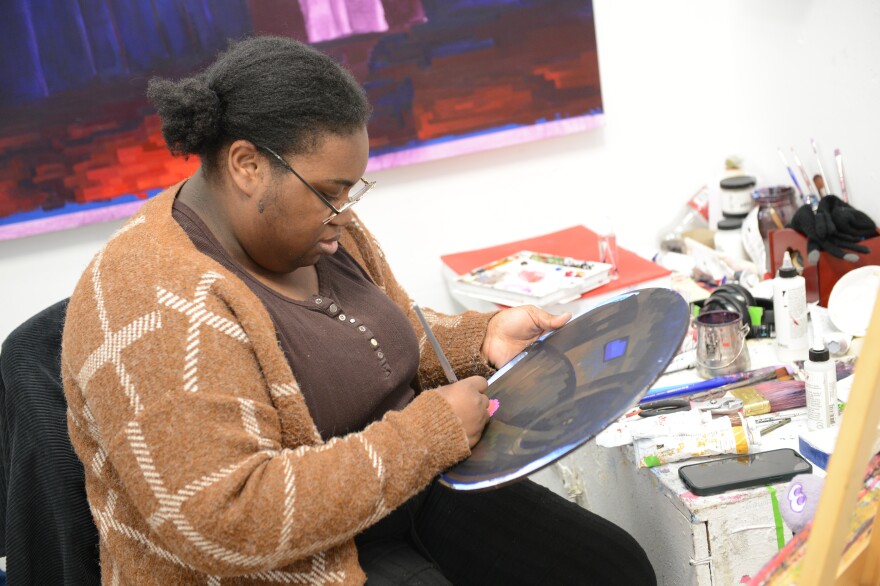 Artist painting on small circular canvas in studio filled with brushes and paint.