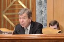 Close-up of US Sen Roy Blunt seated during a Senate committee meeting. He is wearing a dark suit, white shirt and silver patterned tie with a microphone and his nameplate in front of him.