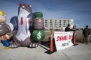 Three men can be seen standing outdoors on a corner of an intersection. They are standing behind a banner that reads "Shame on Port KC." At left is a large inflatable cat that is is smoking a cigar wearing a suite. It  has its paws wrapped around an inflatable person who looks like a construction worker in its right paw and a bag of money in its left paw.