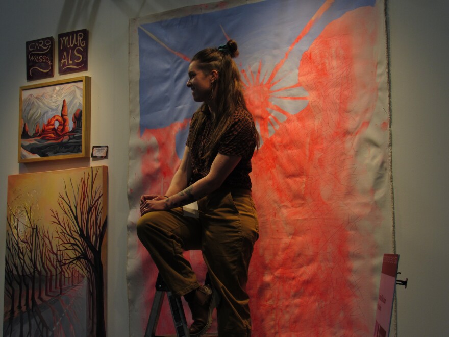 A woman sits on the top rung of a ladder in front of a large canvas painting
