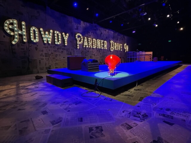 A blue raised platform sits beneath a sign reading howdy pardner drive in in bright lights.