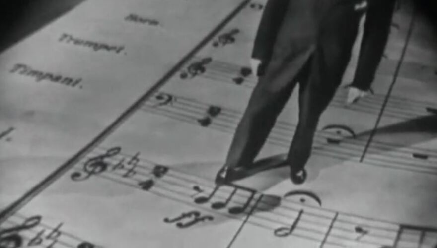 black-and-white photo of Leonard Bernstein pointing with his foot to notes on an oversized score of Beethoven's Fifth Symphony