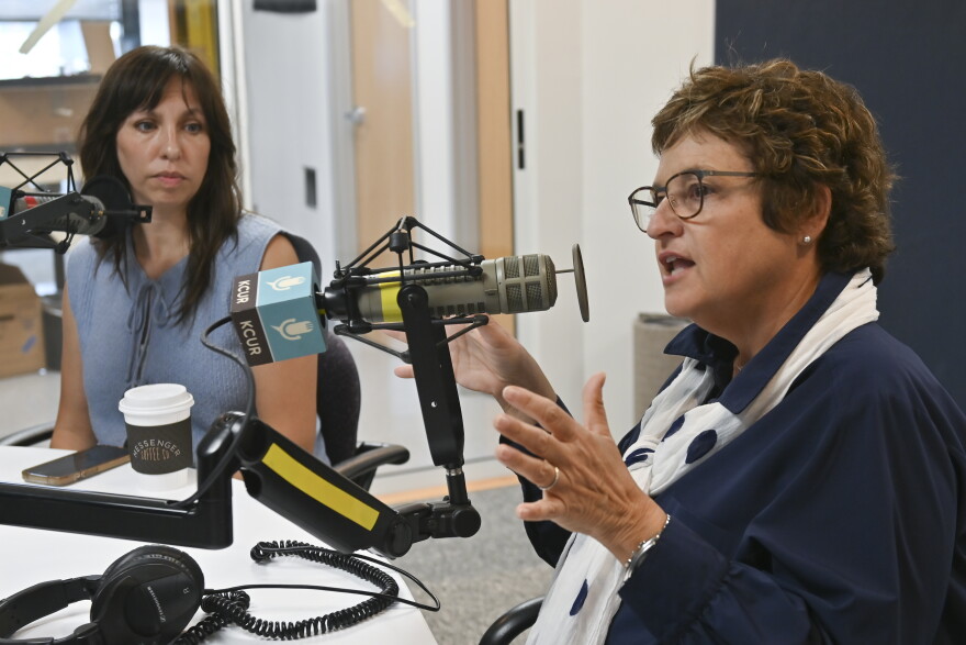 Two people sit in a radio studio talking at microphones. The person at right, Sarah Morris, is gesturing while talking.