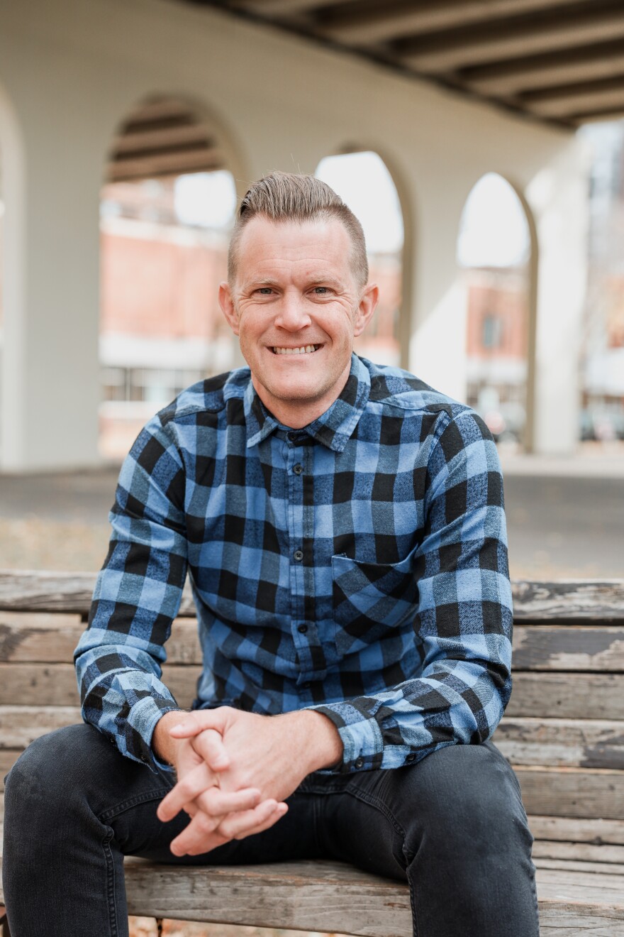 A man sitting on a wooden bunch with a blue and black pattern shirt.