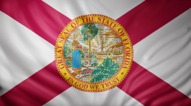 Renderizado en 3D de una detallada bandera de Florida, de color blanco, con franjas rojas que descienden desde las esquinas formando un patrón en forma de X. El sello situado en el centro presenta un borde exterior amarillo y lleva inscritas las palabras: "Great Seal of the State of Florida" y "In God We Trust". La ilustración interior muestra a una mujer con una larga falda azul depositando flores rojas y blancas en el agua; al fondo, se aprecian un barco y una palmera.