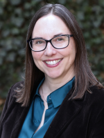 a white woman with dark hair and glasses wearing a teal colored shirt and brown velvet coat smiles and stands in front of a dark ivy background