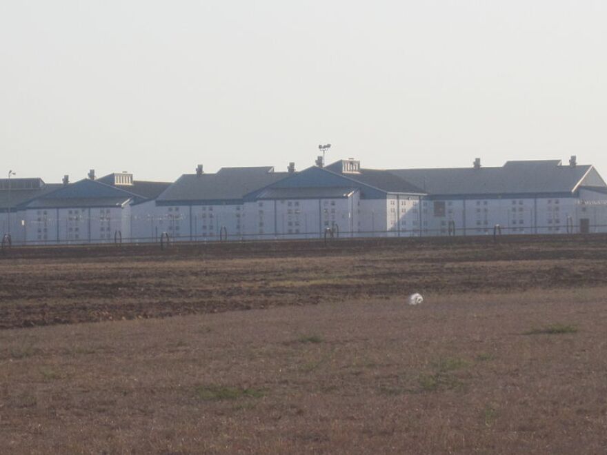  The Dolph Briscoe Unit prison is seen on the horizon. It's a long, gray building dotted with tiny windows and surrounded by fences. A brown grassy field is in the foreground.