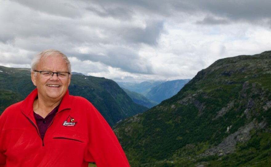 A man with a red fleece stands in front of Norwegian mountains with clouds above.