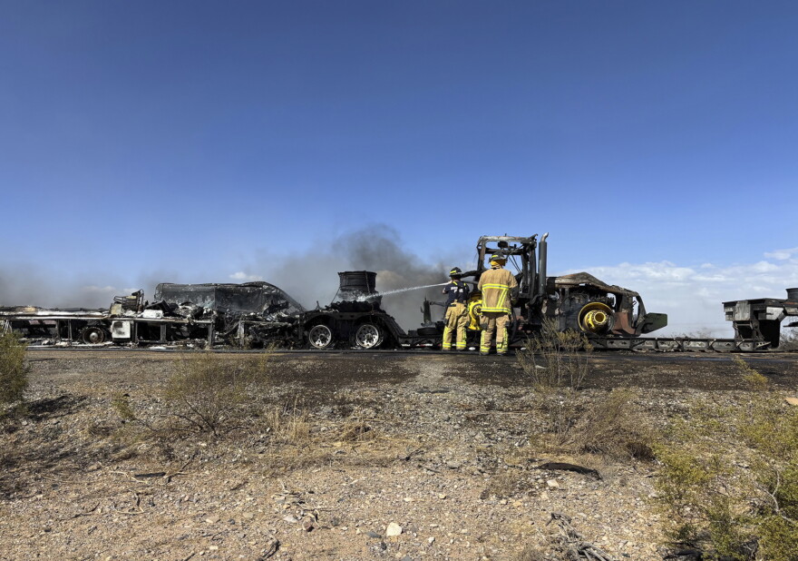 Firefighters hose water on burnt tractor-trailers and cars enveloped in smoke on the side of a rural, desert highway.