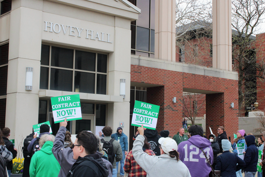 A crowd carrying signs stops at the front of a large brick building, with 'HOVEY HALL' denoting the building name.