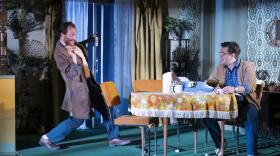 Man in coat stands by table where man sits