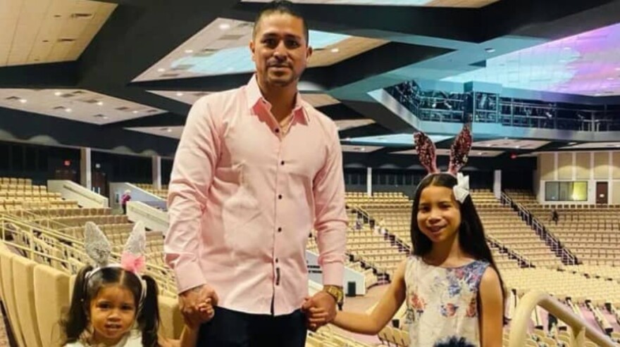 A man in a beige button-down shirt holding two girls' hands on either side while standing in an arena