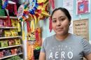 A young Hispanic woman stands in her party shop with a bible verse in the background