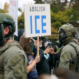 Anti-ICE protestors stand facing police officers who are wearing gas masks and camouflage fatigues.