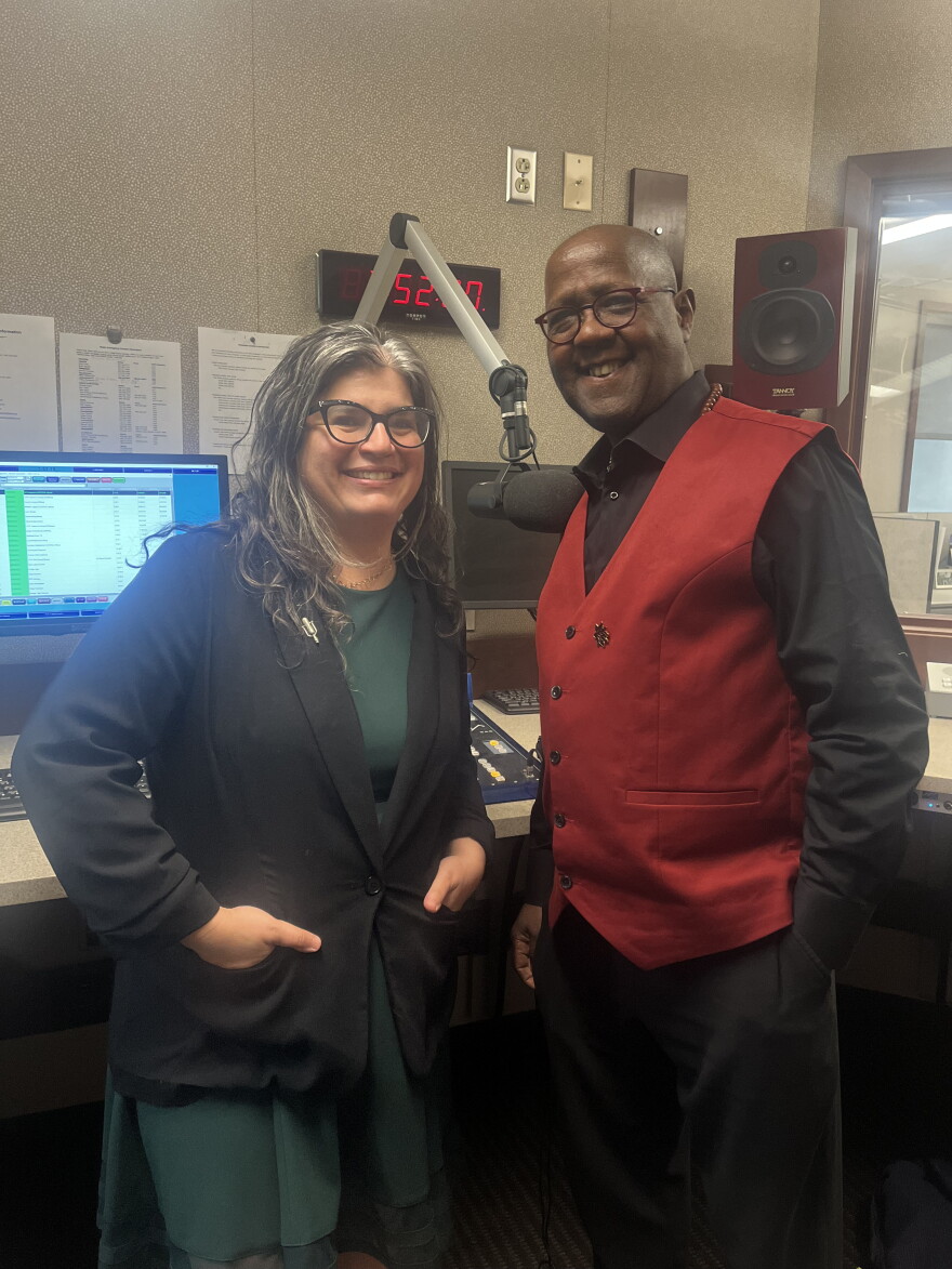 On the left, Mona Seghatoleslami smiling wearing a green dress and black jacket. On the right, viola professor George Taylor, also smiling, wearing a black shirt with a red vest. They are standing in a studio in front of a microphone and a computer.
