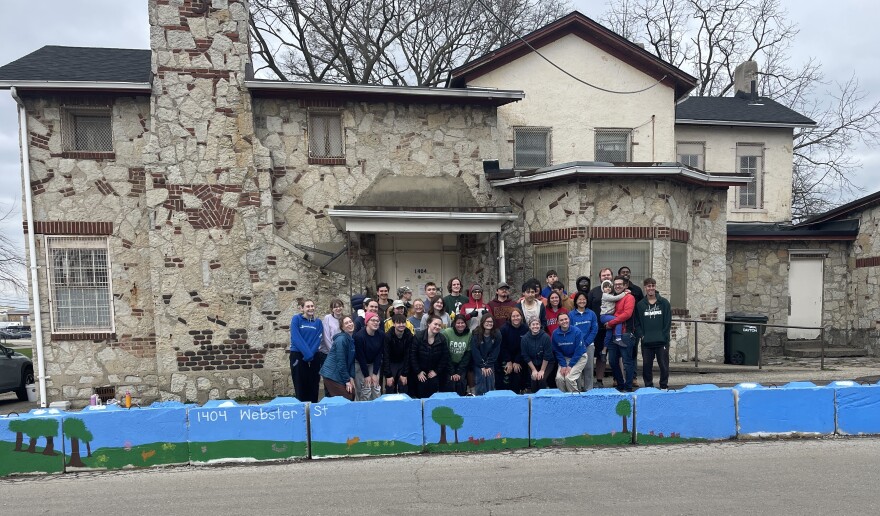 A large group of people stand in front of a large stone house, with a mural of a green hill and trees and a blue sky on a stone wall.