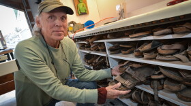A man wearing a cap and long-sleeve pullover with glasses on a strap that dangle from his neck pulls out a worn pair of Birkenstock sandals from a shelf full of other worn, used Birkenstock sandals.