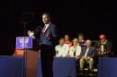 man stands at a microphone. Trump/Vance signs are next to him and six people are seen seated behind him.