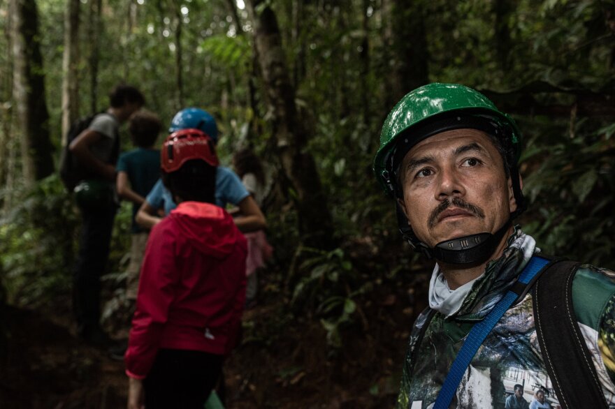 A man wearing a helmet and outdoor gear looks in the distance while facing the camera, as a line of people wearing raincoats and helmets walk through the jungle behind him.