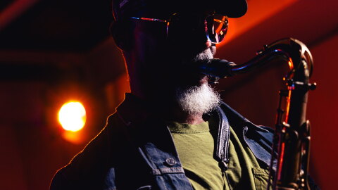 A man inside a recording studio. His face is in shadow, the light illuminating the rims of his glasses as he plays the saxophone. 