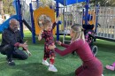 A young toddler walks from one adult toward another on a playground