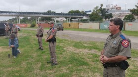 Jesse Fuentes kneels near the Rio Grande while surrounded by Texas DPS troopers while in front of a wall of cargo cantainers and razor wire blocking access to the river.