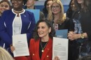 Governor Gretchen Whitmer sits at a desk and holds up two printed documents. She smiles. She wears a bright red blazer with gold buttons.