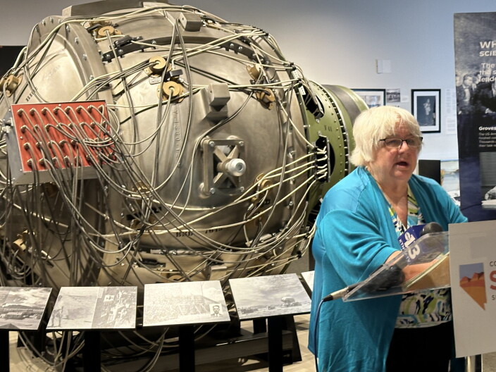 A white-haired, bespectacled woman speaks at a podium. Behind her is a large metal contraption with wires winding around all over. It's a replica of an atom bomb.