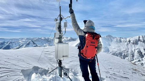 A person with a backpack stands on a snowy mountain and attends to a device.