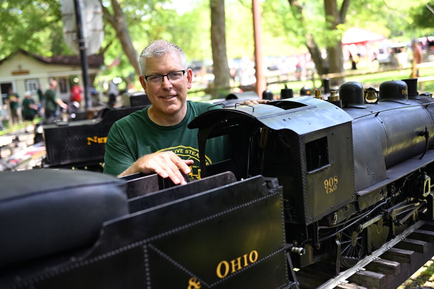 A man poses outdoors next to a miniature steam engine
