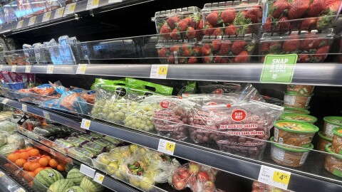 Interior photo of a grocery store refrigerated section that is open-air. A variety of fruits and vegetables can be seen packed on the shelves.