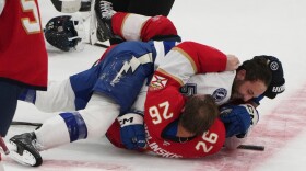 A hockey player in blue and white is on top of an opponent in red, white, and black. They're both trying to punch each other, as the helmet on the player on top is falling off his head.