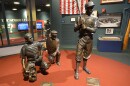 Two bronze statues of baseball players and an umpire stand on a fake dirt infield that resembles home plate inside the Negro Leagues Baseball Museum. Behind them is an American flag and a black and white photo of a baseball team. 