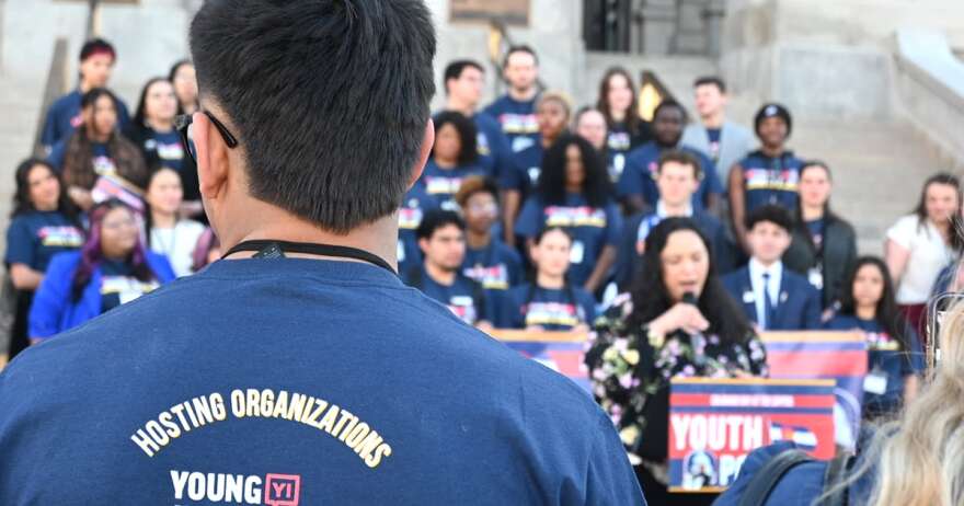 A large crowd stands behind a woman at a podium.