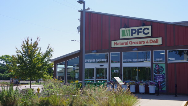 a red building with a slanted roof sits in front of thin flowers, weeds and other plants on a concrete walking path. The sign for the store reads "PFC Natural Grocery & Deli."